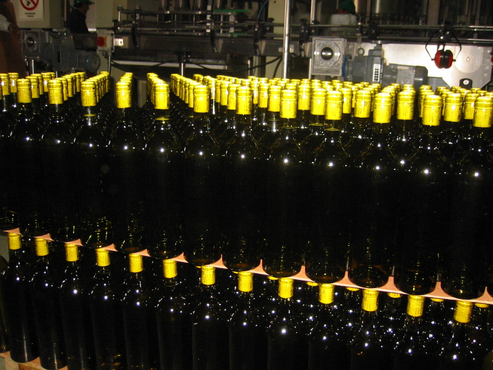 Many dark wine bottles with yellow caps stacked in rows in a bottling facility.