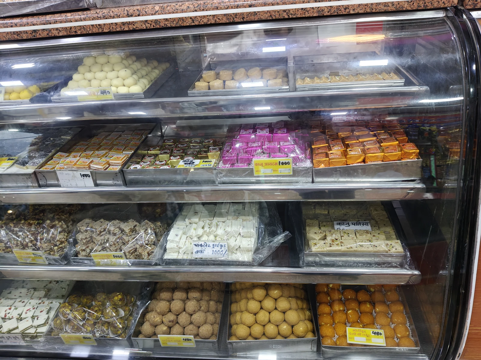A variety of Indian sweets are displayed in a refrigerated glass case.