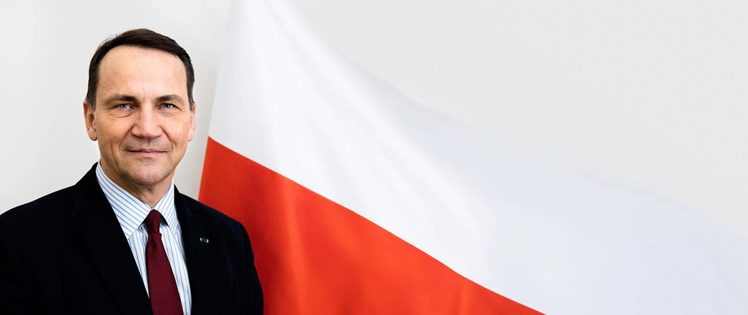Radosław Sikorski, Polish Minister of Foreign Affairs, smiling in front of a Polish flag.