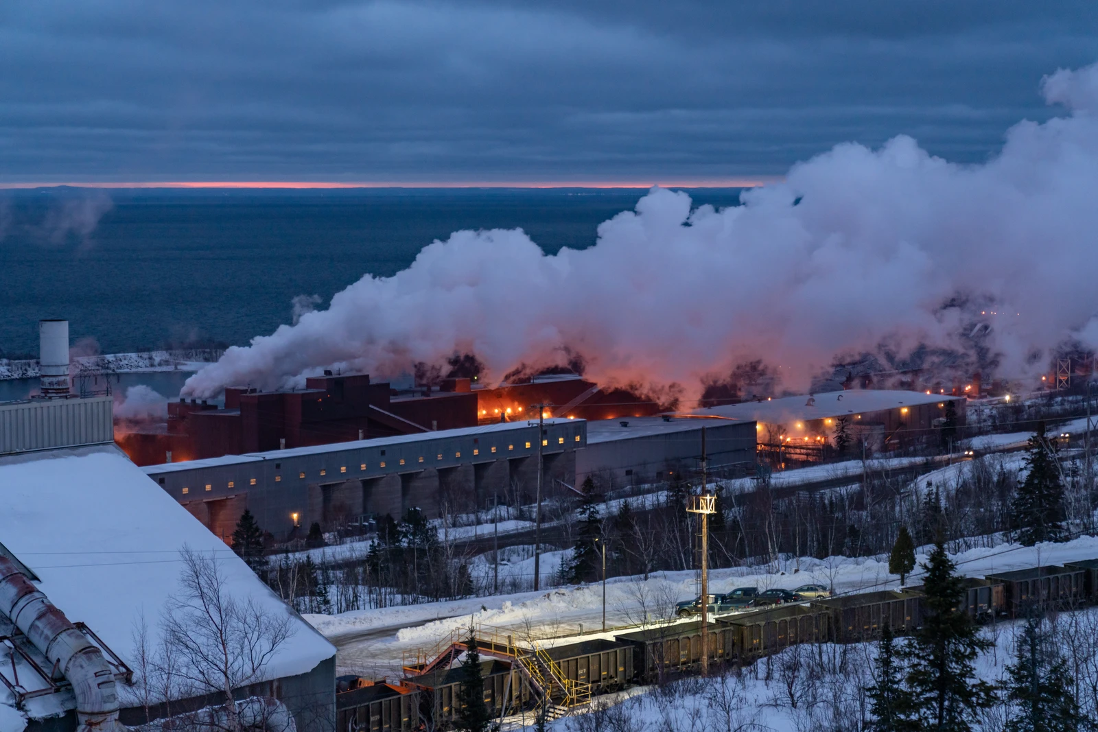 Iron ore mining plant with large plumes of steam or smoke rising into the evening sky.