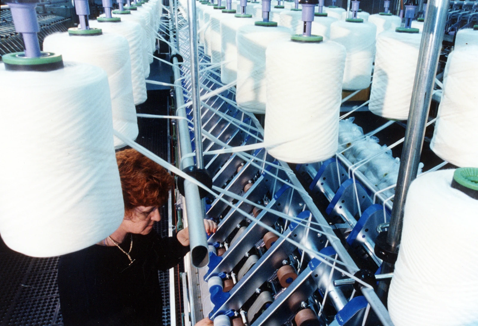 Woman working with textile machinery that processes white spools of yarn.