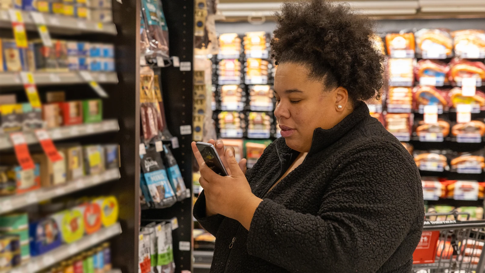 Woman looking at a smartphone while standing in a grocery store aisle.