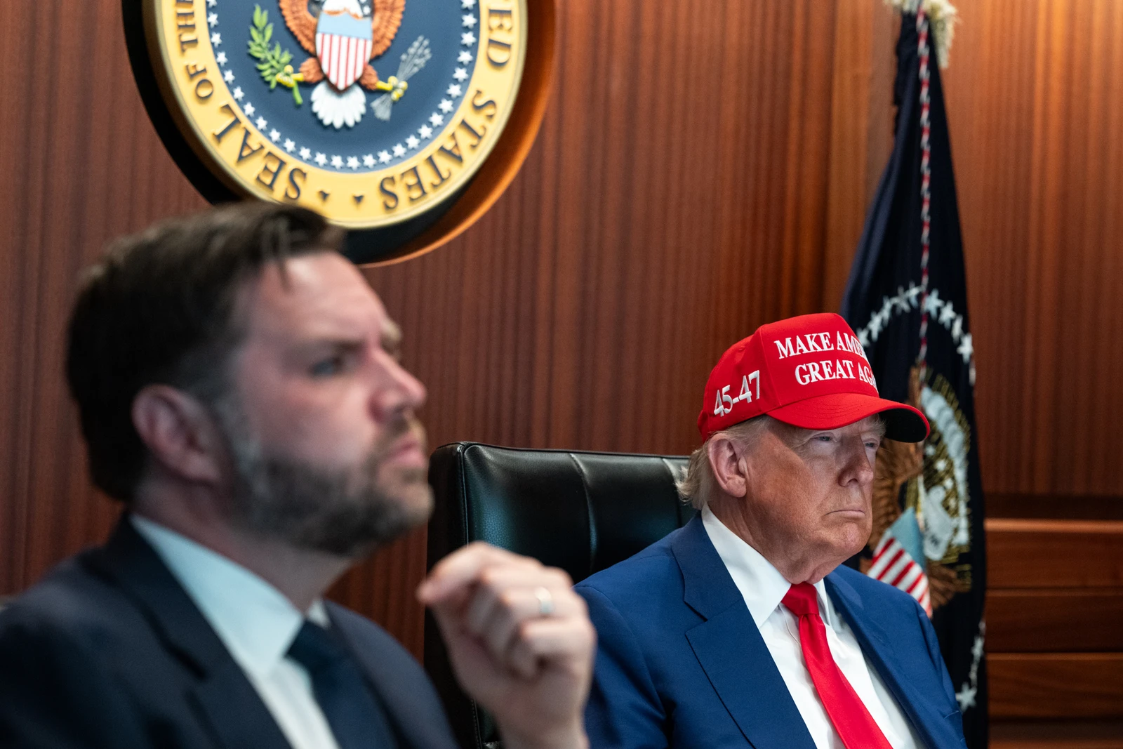 Former President Donald Trump seated during a meeting, wearing a red hat.