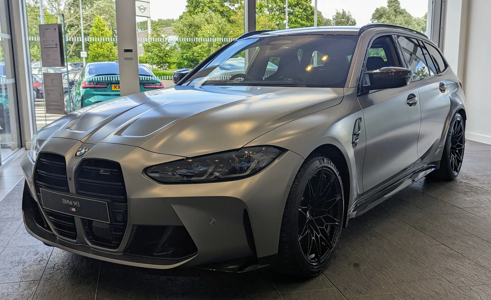 A silver 2023 BMW M3 Touring parked inside a car dealership.