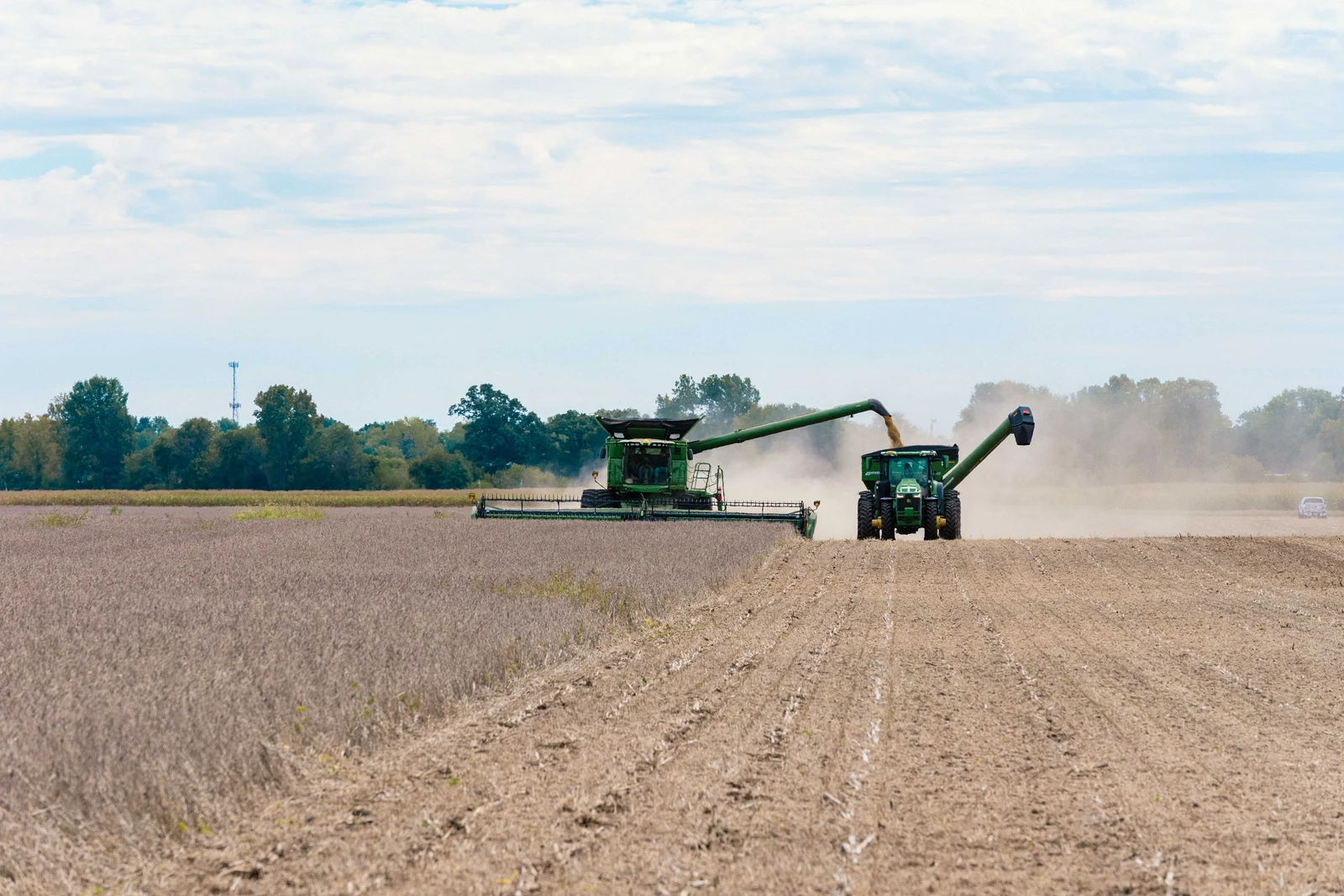 Combine harvester on the left moving through a field, pouring soybeans into a tractor on the right.